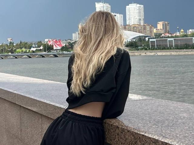 LauraleePenderel with long, wavy blonde hair, black top, and black skirt, leaning on a concrete wall, facing a waterfront, city skyline in the background, overcast sky.