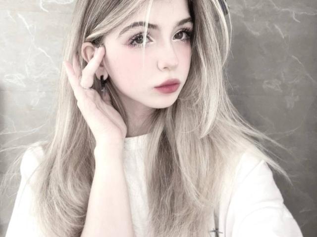 LauraleePenderel with long blonde hair, pale skin, and pink lips, wearing a white shirt, hand touching her ear, looking slightly to the side, with a soft, dreamy expression.
