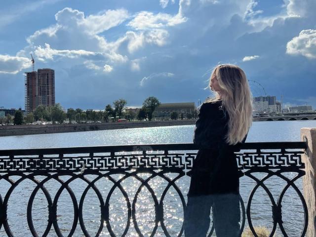 LauraleePenderel with long blonde hair, black top, and blue jeans stands by a sparkling river, facing left, against a bright sky and city skyline with a Ferris wheel.