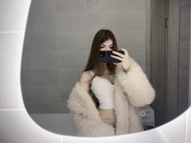 LauraleePenderel with long brown hair, white crop top, and fluffy beige coat, holding phone in front of mirror, showing her tits and a small tattoo on her right hip.