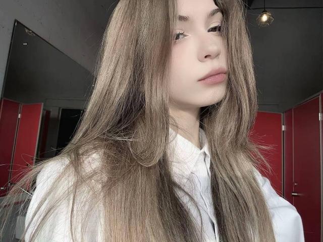 LauraleePenderel with long, straight brown hair, white shirt, and a neutral expression, standing in a room with red doors, has pale skin and a slight pout on her lips.