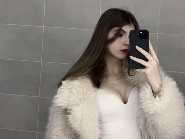 LauraleePenderel with long brown hair, white dress, and fluffy beige coat, holding a black phone, showing her tits, red lipstick, and a necklace, in a mirror selfie.
