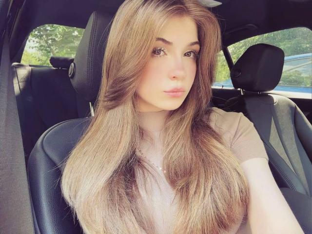 LauraleePenderel, sitting in a car with long, straight brown hair, wearing a beige top, looking slightly to the side with a neutral expression. Boobs not visible.
