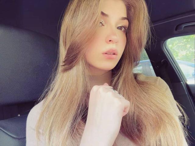 LauraleePenderel has long, straight, blonde hair, pale skin, and pink lips. She's touching her neck with one hand, wearing a light pink top, and has a small nose piercing.