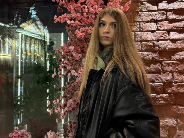 LauraleePenderel with long brown hair, green scarf, black leather jacket, standing against a red brick wall with pink cherry blossom branches in the background. She looks serious.