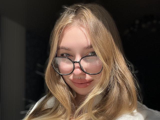 DaniellaDella, mit blonden Haaren, trägt eine Brille, hat ein Nasenpiercing und lächelt mit rosafarbenen Lippen. Sie trägt ein weißes Oberteil und ist im Sonnenlicht. Ihr Hintern ist nicht sichtbar..