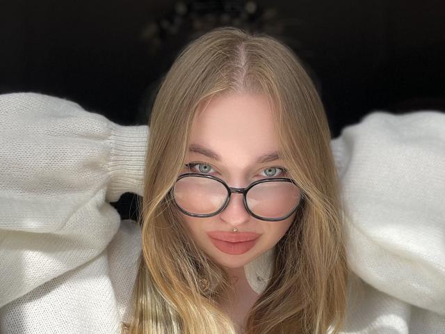 DaniellaDella mit blonden Haaren, grünen Augen und einer Brille mit schwarzem Rahmen, trägt einen weißen, flauschigen Pullover, hat die Hände im Haar, lächelt mit einem Nasenpiercing und hat volle, rosafarbene Lippen..