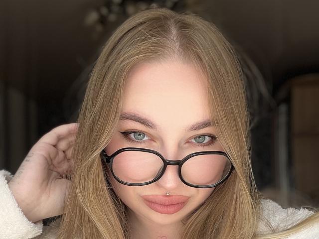 DaniellaDella, con i capelli biondi, gli occhiali neri e un piercing al naso, guarda la fotocamera con gli occhi azzurri e le labbra rosa, indossando un maglione bianco e soffice..