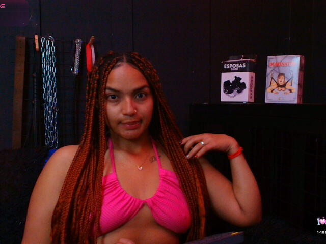 BrianaPratt with long red braids, wearing a pink bikini top, showing her tits, nose ring, and red bracelet, in a dimly lit room with books and clothes.