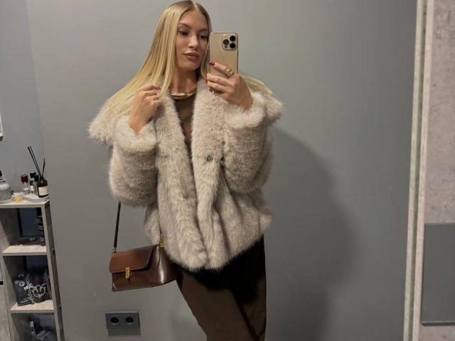 LesliSaberi with long blonde hair, wearing a fluffy grey fur jacket, brown pants, and a brown purse, taking a mirror selfie with a gold phone. Boobs covered by jacket.