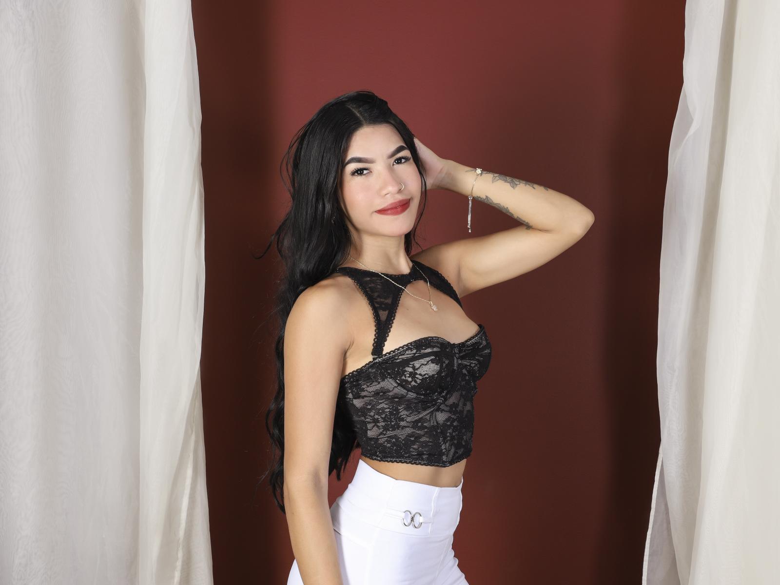Ruby Castillo, com cabelos longos e pretos, vestindo uma blusa cropped de renda preta e calças brancas de cintura alta, sorrindo, com a mão no cabelo, tatuagem no braço direito, fundo vermelho, cortinas brancas..