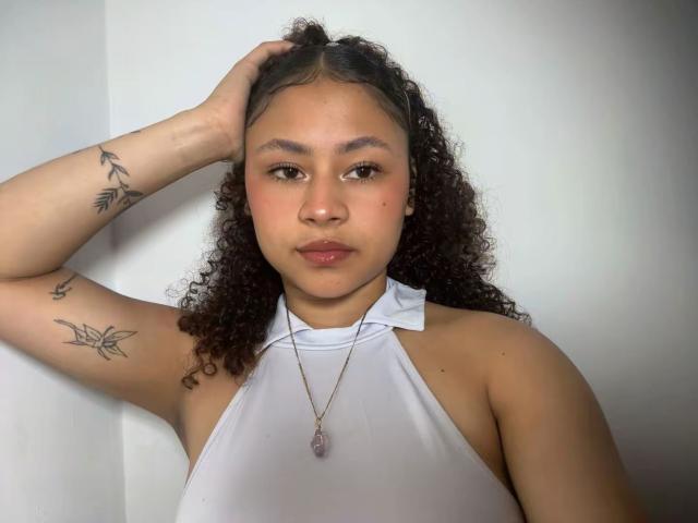 SofiaPassionne with curly dark hair, brown eyes, and light brown skin, wearing a white halter top, hand in hair, tattoos on arm, necklace with pink stone, neutral expression.