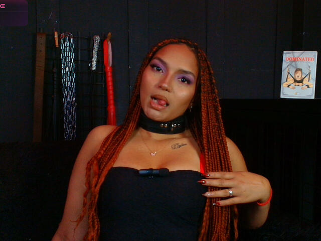 BrianaPratt with long red braids, purple eyeshadow, black choker, and black strapless top, tongue out, hand on chest, showing small tits, ring on finger, dark background.