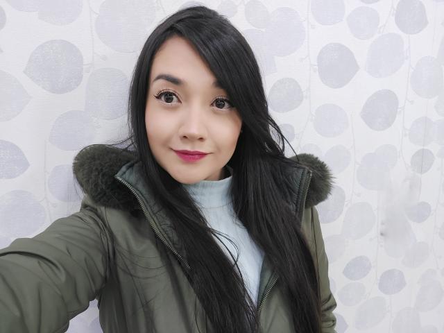 Annhy, con cabello negro largo, una chaqueta verde y una capucha forrada de piel, sonriendo a la cámara, mostrando lápiz labial rosa y un rostro adorable, vistiendo un cuello alto azul claro..