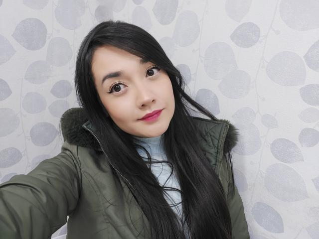 Annhy, met lang zwart haar, een groene jas en roze lippenstift, kijkt naar de camera en toont haar gezicht en bovenlichaam, terwijl ze een lichtblauwe coltrui draagt..