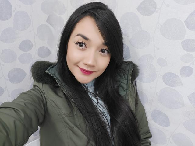 Annhy, con i suoi lunghi capelli neri, la giacca verde e il rossetto rosa, sorride alla fotocamera, mostrando il suo viso carino e il suo piccolo naso. I suoi occhi marroni sono concentrati..