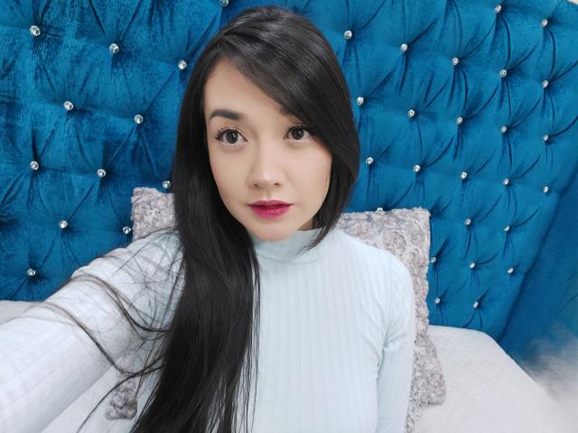 Annhy with long black hair, light blue ribbed shirt, pink lipstick, and serious expression, sitting against a blue, tufted, diamond-studded headboard. Her tits are small and her pussy is unseen.
