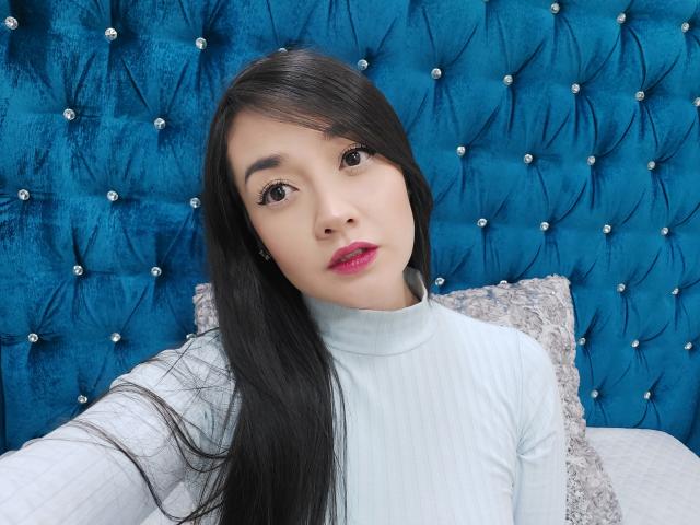 Annhy a de longs cheveux noirs, une peau claire, du rouge à lèvres rose, et porte un pull col roulé blanc texturé. Elle est assise devant un tête de lit bleu en velours texturé, orné de clous en forme de diamants..