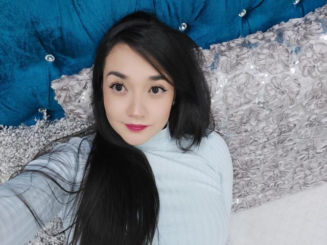 Annhy, com cabelos longos e pretos, pele clara e batom rosa, vestindo um suéter azul claro, deitada em uma cama azul com detalhes em relevo e um travesseiro prateado com lantejoulas, tirando uma selfie..