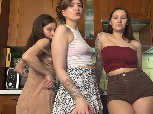 Three HotCaramelsTrio standing in a kitchen, wearing casual clothes: one in a pink dress, one in a white tank top and floral skirt, one in a red strap top and brown shorts.