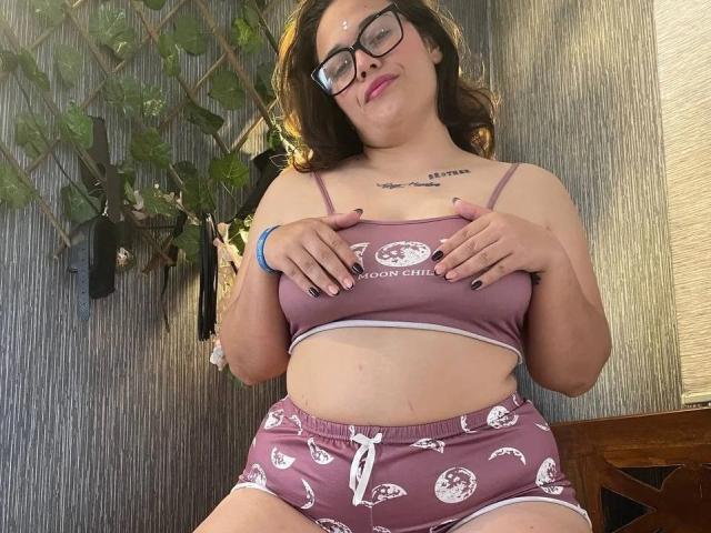 BBWCouple, hard uiterlijk, met een bril, middelgrote borsten, bruin haar, draagt een paars sportbeha en bijpassende korte broek, handen op de borst, lachend, paarse nagels, tatoeage op de borst, zonlicht, houten muur..