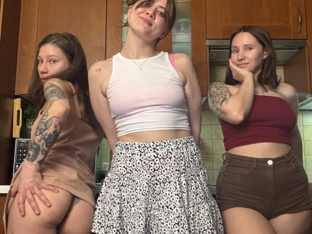 Three HotCaramelsTrio standing in a kitchen, wearing tank tops and shorts, exposing their asses. One has tattoos, one has a pink bra, all have cute smiles.
