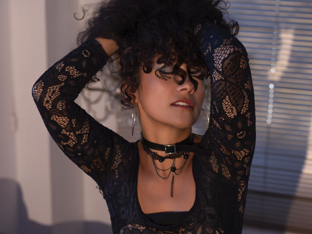 SugerVibes with curly black hair, wearing a black lace top, choker, and earrings, arms raised, looking seductive, sunlight on face, medium skin, slightly parted lips, indoor setting.