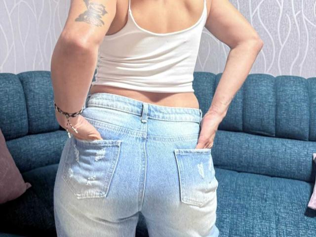 SharonBellarie ha vuelto, vestida con una camiseta blanca y unos vaqueros azul claro, ligeramente rotos, con las manos en los bolsillos, mostrando un tatuaje en su brazo derecho, sentada en un sofá azul..