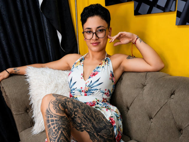 ConnieJonnes, con i capelli neri corti, gli occhiali, un vestito floreale e tatuaggi sulla coscia, seduta su un divano grigio, sorridente, con un braccio alzato e un cuscino bianco e soffice alla sua sinistra..