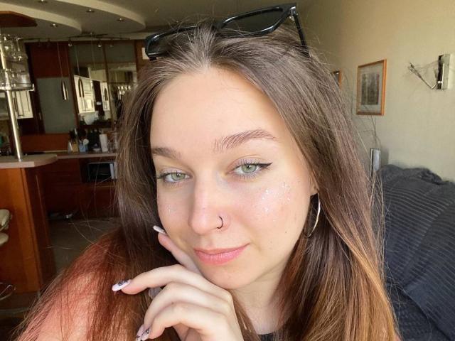 ReynaGueningsman has long brown hair, green eyes, a nose ring, and is wearing hoop earrings. She's touching her chin with a slight smile. Sunglasses rest on her head.
