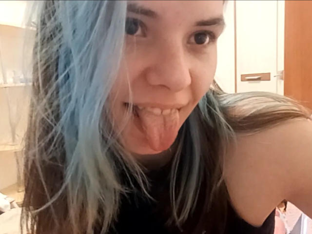 MathaDernier mit blau gefärbten Haaren, die Zunge herausstreckt, in die Kamera schaut, kleine Zähne zeigt, ein schwarzes Hemd trägt, in einem Selfie, hat helle Haut und dunkle Augenbrauen..