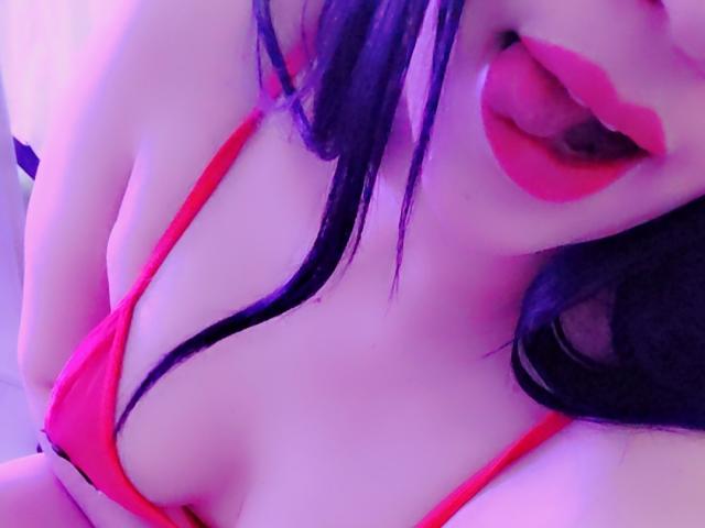 ValeriaRossii with purple-black hair, red lips, and tongue out, wearing a red bra that shows her tits, under purple-blue lighting.