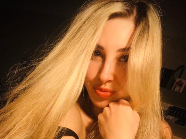 Blonde VelvetObsession with long hair, orange lipstick, and black top, resting chin on hand, smiling softly, warm light on face, slightly messy hair, looking at camera.