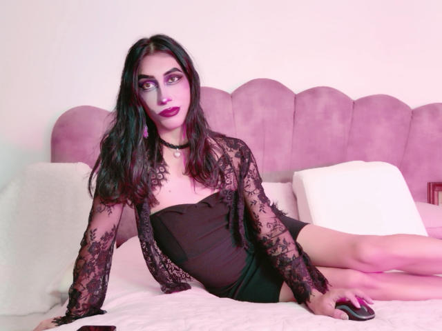 AlexaMissSx, avec un rouge à lèvres violet foncé, un haut en dentelle noire et un short noir, assise sur un lit, les jambes croisées, portant un collier noir, et regardant la caméra..
