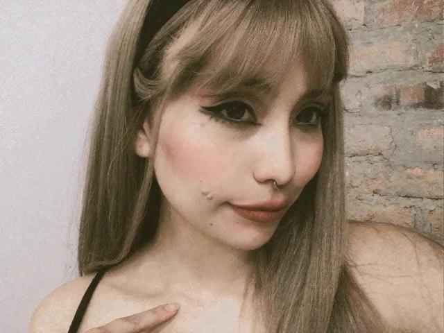 Ilayna has long brown hair, black eyeliner, and a nose ring. She's smiling with her hand on her chest, showing a small mole on her left cheek.