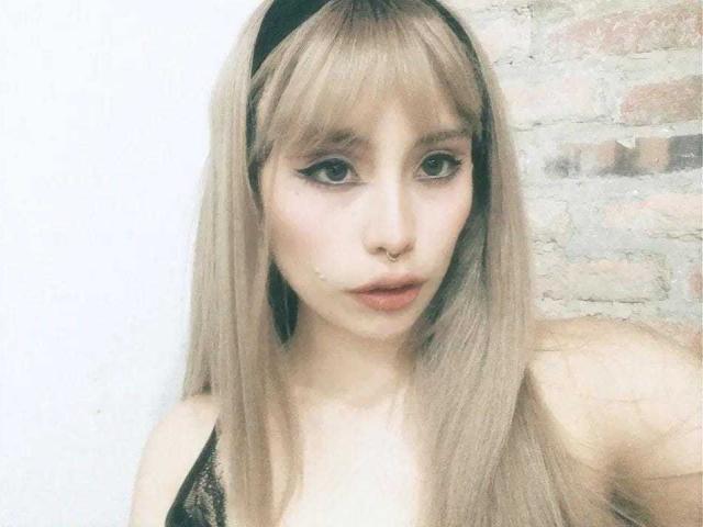 Ilayna has long blonde hair, a black headband, and a nose ring. She's wearing black lace, with winged eyeliner and parted lips. Her tits are small.