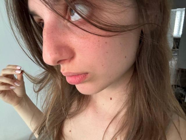Le visage de KarieMestler, avec ses joues rosâtres, ses taches de rousseur et une petite mole près de son nez. Elle tient ses cheveux bruns avec ses doigts aux ongles roses, et a un léger air boudeur..