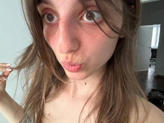 KarieMestler ha labbra rosa e carnose, occhi marroni e un viso leggermente arrossato con delle lentiggini. Sta facendo un'espressione accigliata, tenendosi i capelli castani con una mano..