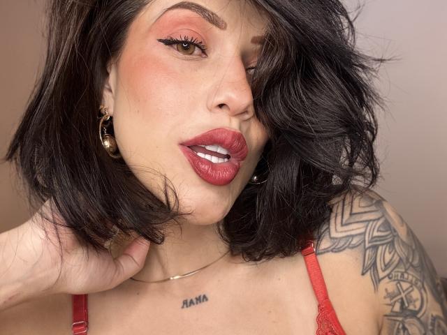 HannaBorbon, com cabelo castanho ondulado, batom vermelho, brincos de ouro, braço tatuado e sutiã vermelho, segurando o pescoço, olhos castanhos, maquiagem discreta e uma expressão sexy e confiante..