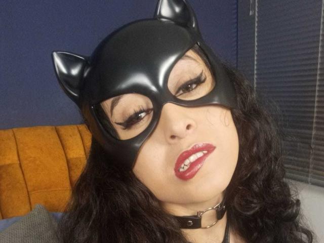 KendraSimons, avec un masque de chat noir, des cheveux noirs bouclés, du rouge à lèvres rouge et un collier noir, regardant vers le haut avec les yeux à moitié fermés, montrant ses dents. Ses seins sont partiellement visibles, et son visage est centré..