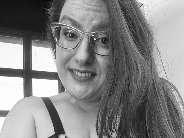 Ein Schwarz-Weiß-Selfie von BadAngel mit Brille, langem Haar, einem Lächeln, einem Trägerkleid, das ein wenig Ausschnitt zeigt, und einem kleinen Nasenpiercing auf der rechten Seite..