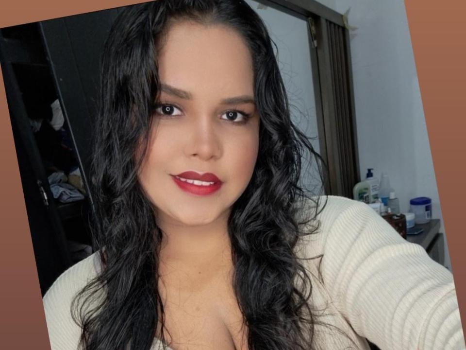 MillySofia, con el cabello negro mojado, lápiz labial rojo y una blusa blanca, aparece en un selfie mostrando ligeramente sus pechos. Está sonriendo y tiene una cara linda..