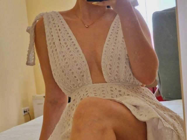 Jumiera indossa un abito bianco con un profondo scollo a V, in pizzo, che mostra il seno e una coscia in un selfie allo specchio, mentre tiene in mano un telefono e indossa una collana d'oro..