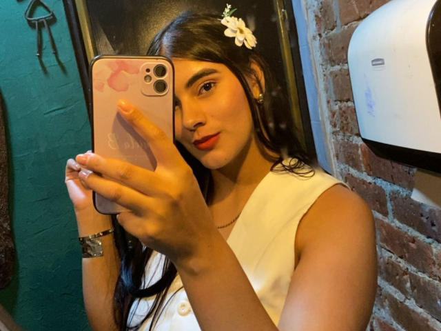 AlaraKey with dark hair, white flower in hair, red lipstick, holding iPhone, wearing white sleeveless top, taking selfie, visible tits, brick wall, green wall, wrist bracelet.