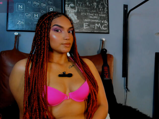 BrianaPratt with red braided hair, pink bra, and a black chest tattoo, sitting with a confident smile, showing her tits, in a room with musical instruments.
