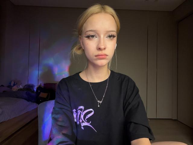 Dallia Banowski, met blond haar, opvallende eyeliner en een neutrale uitdrukking, draagt een zwart shirt met witte graffiti en een zilveren kruisje om haar nek, in een slecht verlichte kamer..