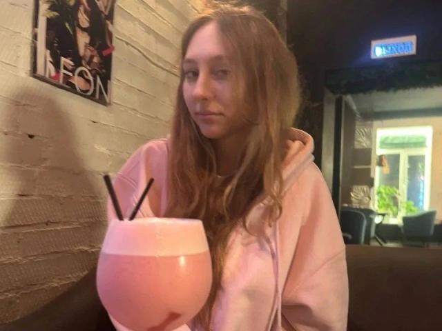 ModelGyrl-hot with long brown hair, wearing a pink hoodie, holding a pink frothy drink with black straws, looking slightly bored. Her tits are small, and she's in a dim room.