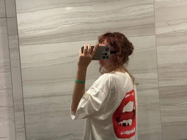ModelGyrl-hot with wavy red hair, wearing a white T-shirt with a red mouth graphic, taking a selfie in a mirror, holding a phone with a green bracelet.