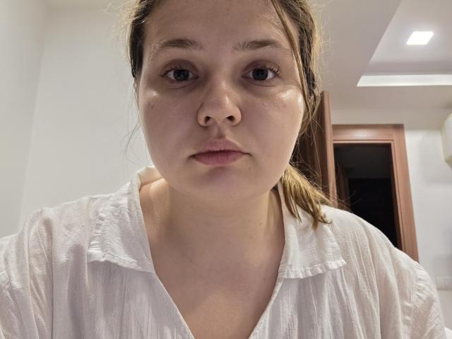 FeSkoczylas hat einen neutralen Gesichtsausdruck, helle Haut, braune Augen und eine kleine Nase. Sie trägt ein weißes, leicht zerknittertes Hemd mit V-Ausschnitt, und ihr braunes Haar ist zurückgebunden..