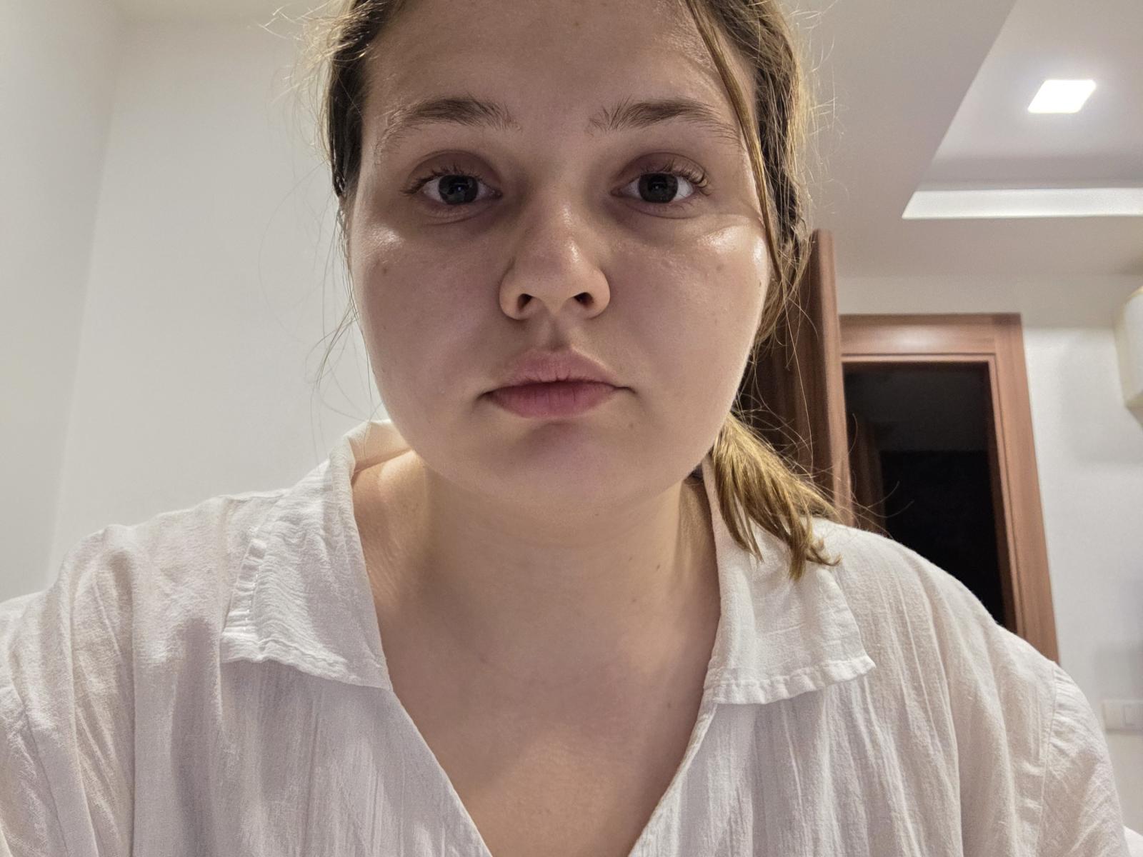 FeSkoczylas has a neutral expression, light skin, brown eyes, and a small nose. She wears a white, slightly wrinkled shirt with a V-neck, and her brown hair is tied back.